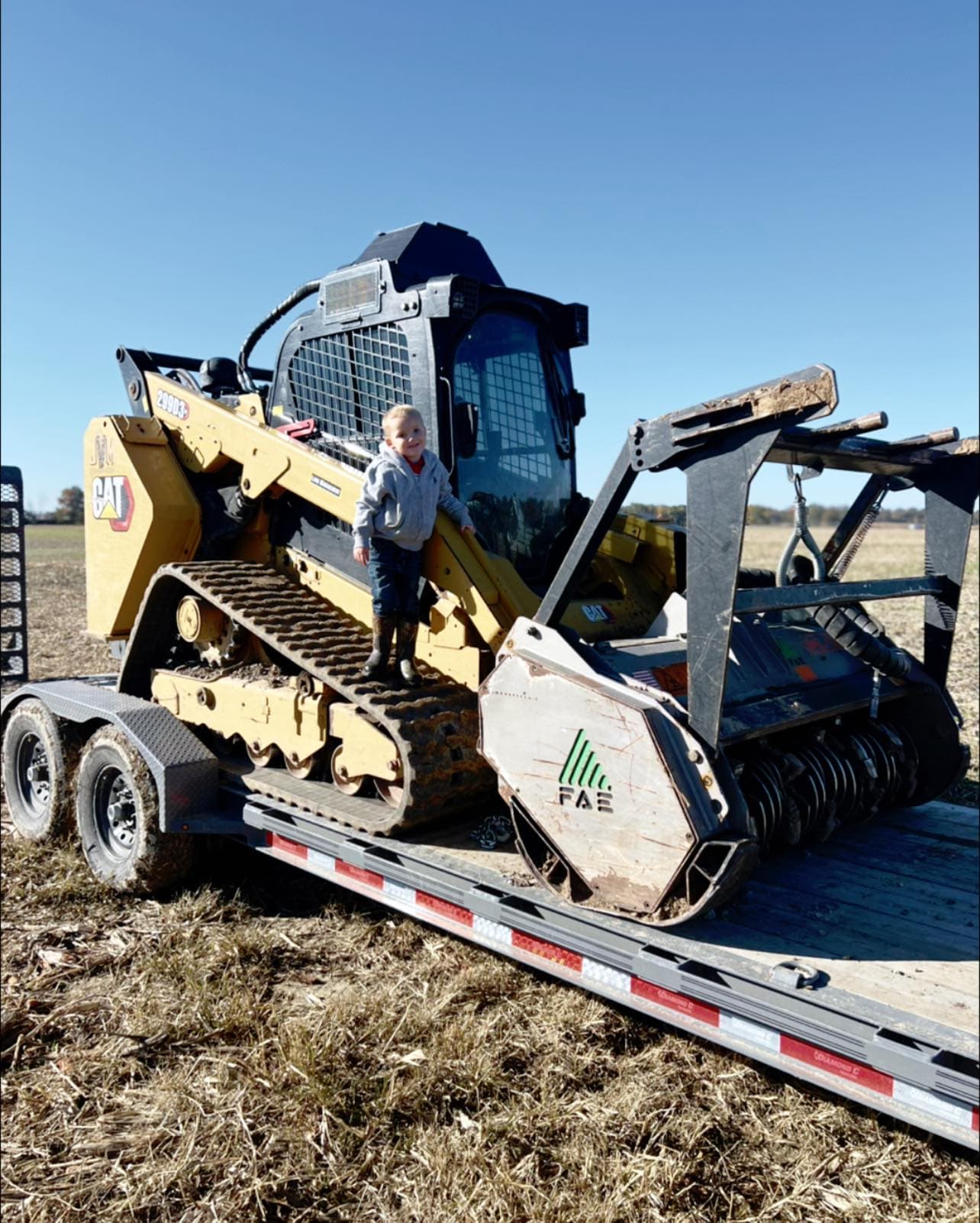 Child playing on a CAT skid steer loader on a trailer in a sunny field.
